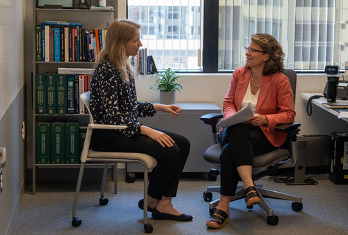 Two scientists sit in an office in front of a bookshelf and window, facing each other.