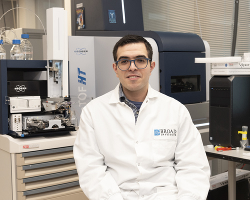 Moe Haines is sitting in a lab wearing a white lab coat. Behind him are various pieces of lab equipment.