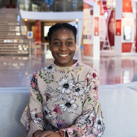 Wolu Chukwu is sitting on a couch in the lobby of the Broad Institute facing the camera and smiling.