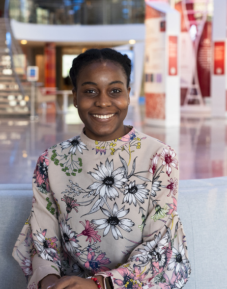 Wolu Chukwu is sitting on a couch in the lobby of the Broad Institute facing the camera and smiling.